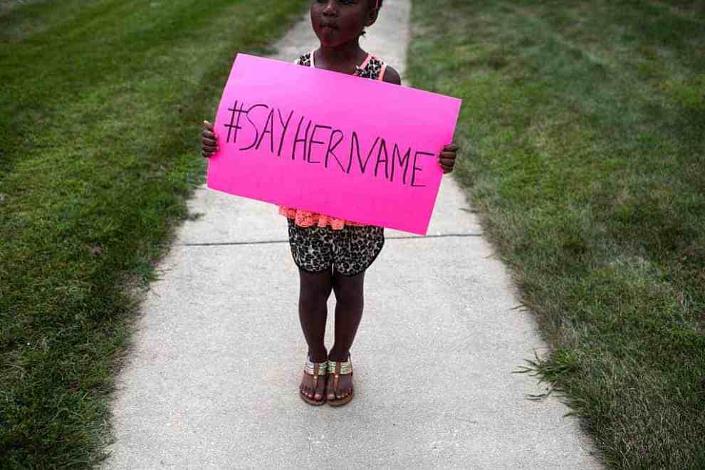 Black girl holding pink sign with black lettering reading #sayhername