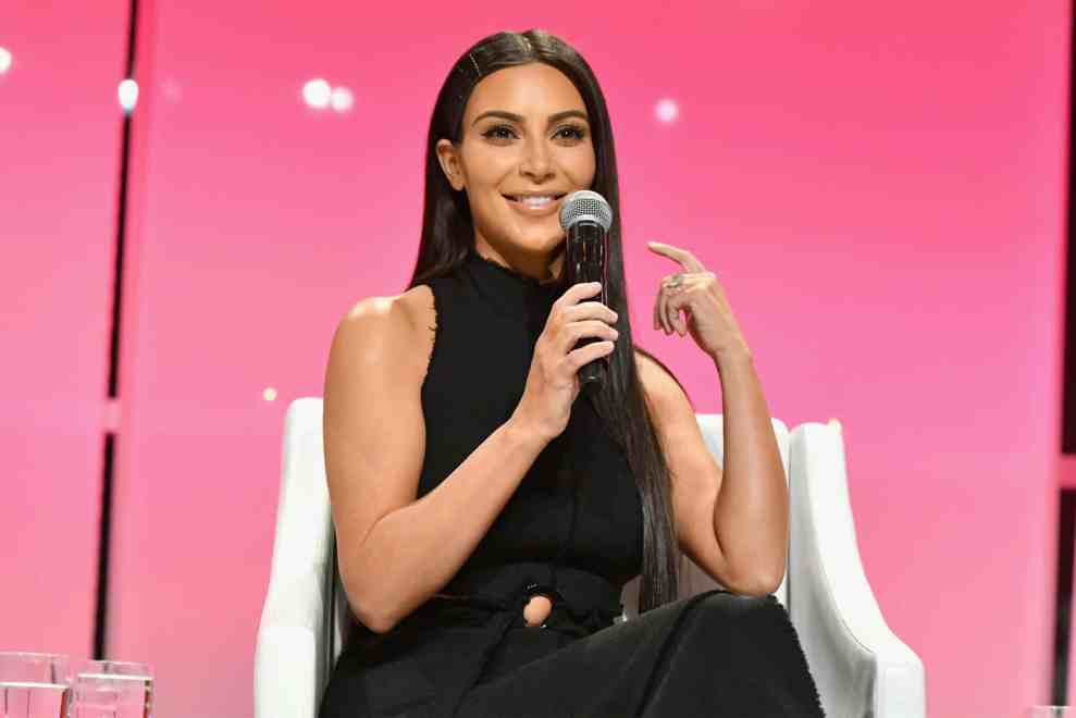 Kim Kardashian in black in front of pink background with microphone