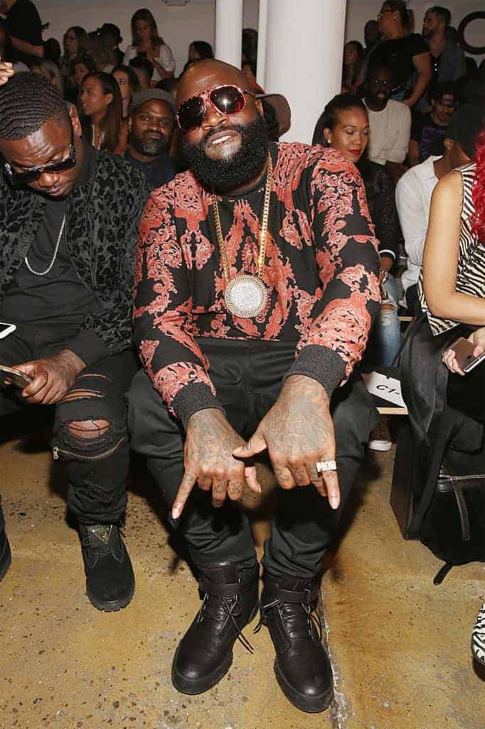 Rick Ross sitting at event wearing black and red jacket with large gold medallion and hands with pinkies out