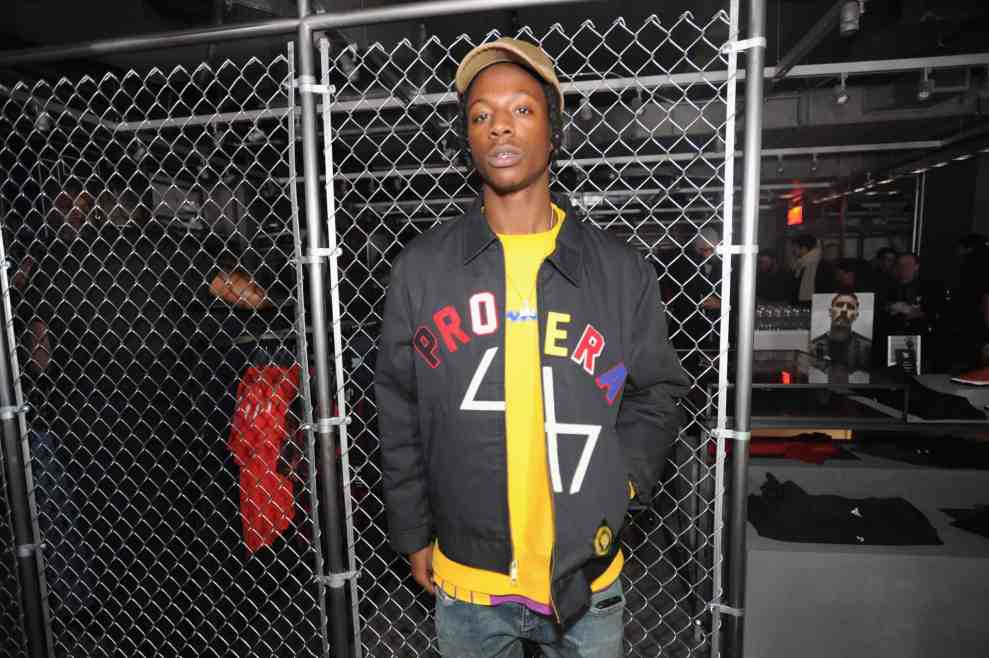 Joey Bada$$ in front of chain link fence wearing Proera 47 zip up hoodie