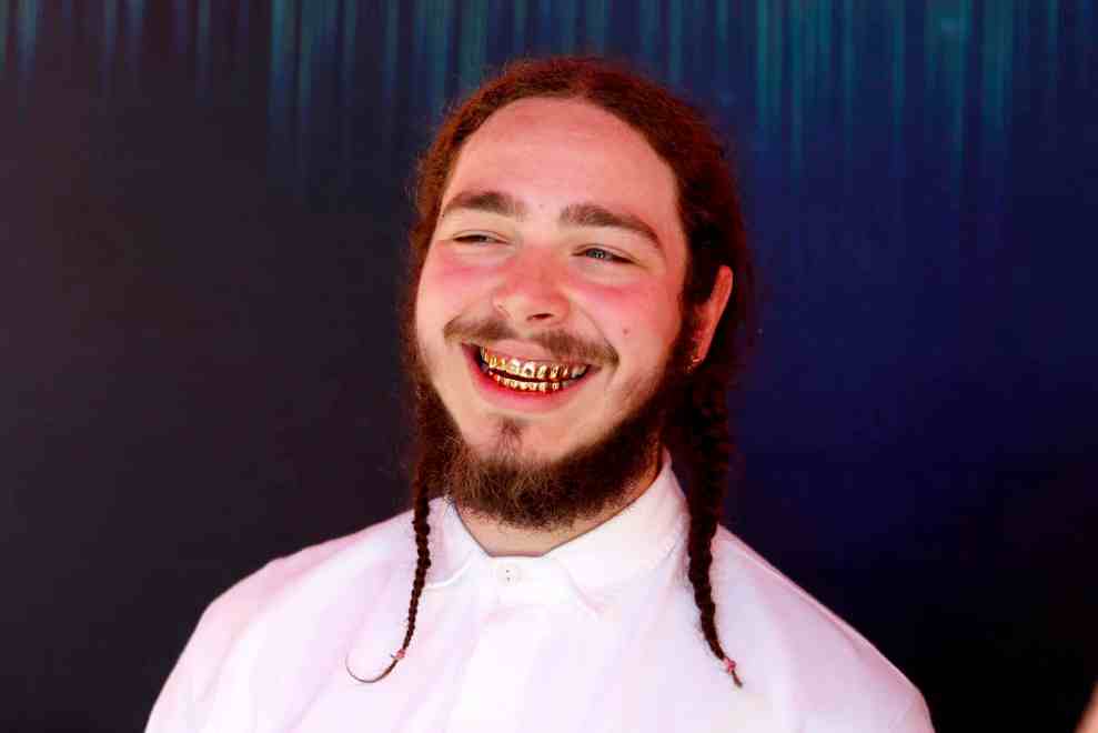 Post Malone with gold grill and two braids in front of blue background