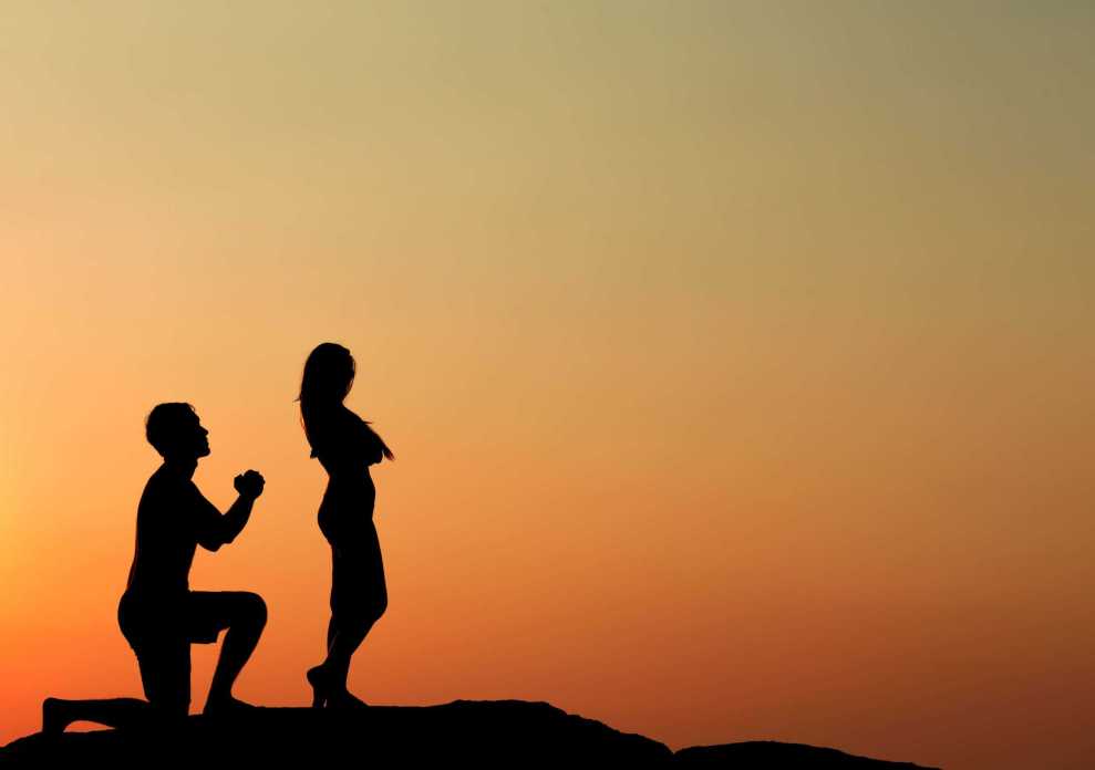 Silhoette of man on one knee with hands clasped and woman facing away from him with arms crossed over her chest at sunset