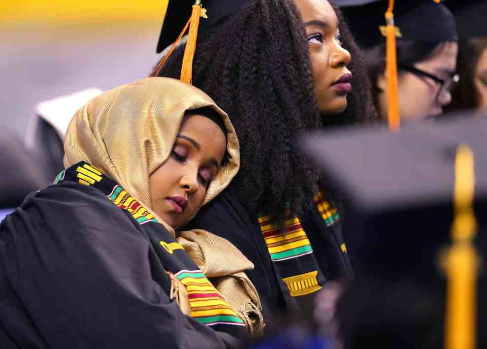 woman in headscarf asleep on another woman's shoulder at a graduation ceremony