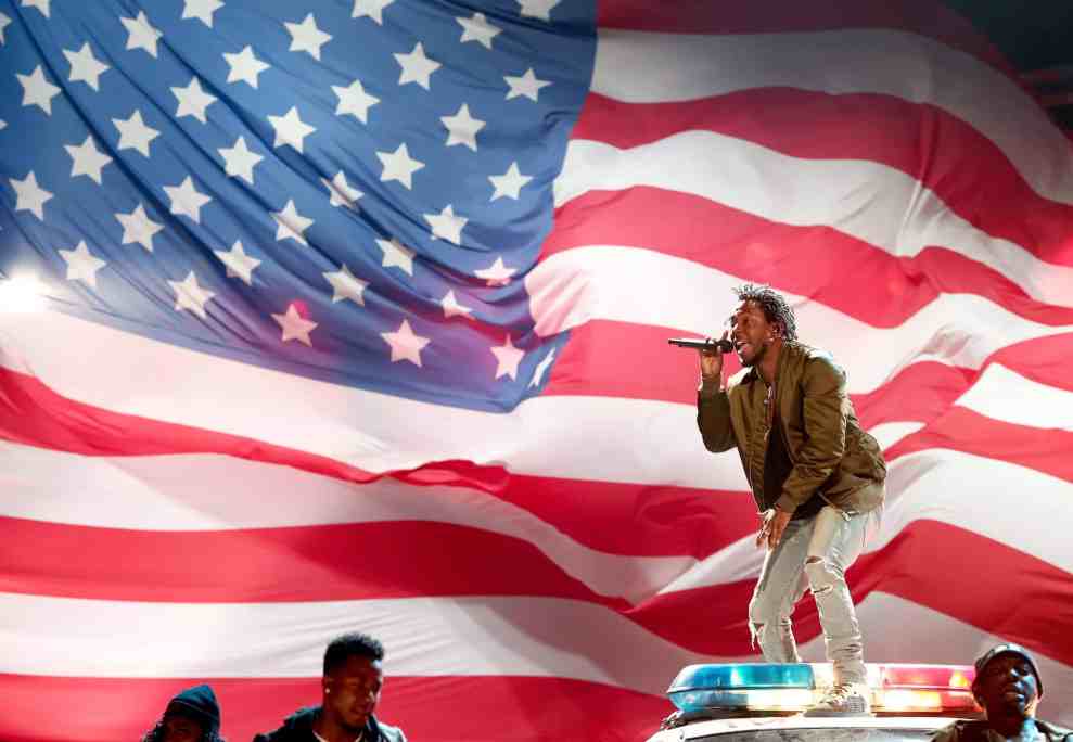 Kendrick Lamar performs on top of police car in front of large American Flag