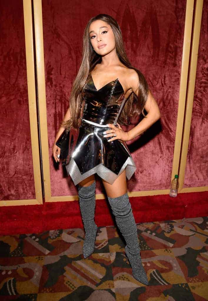 Ariana Grande in short black and silver dress with thigh high silver boots in front of red background