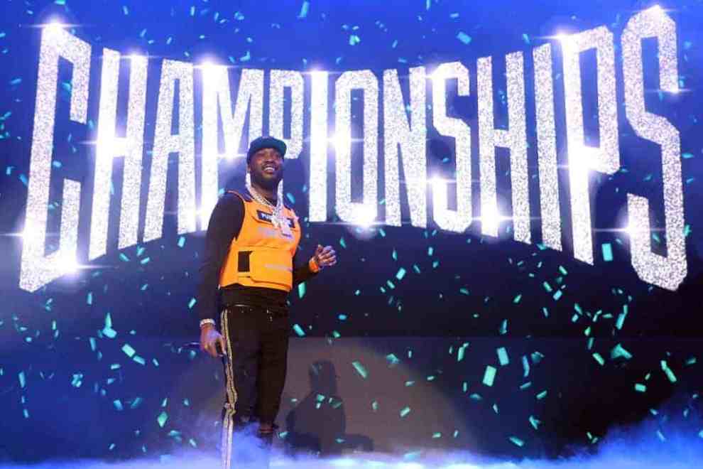 Meek Mill on stage performing wearing a yellow shirt and black shirt standing in front of the Championships sign