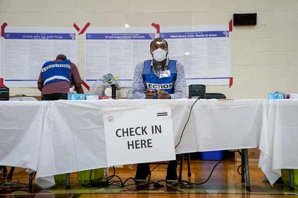 An election official wears a mask and sits behind a plastic barrier as he waits to check in voters at a polling place at McKinley Technology High School during primary election day on June 2