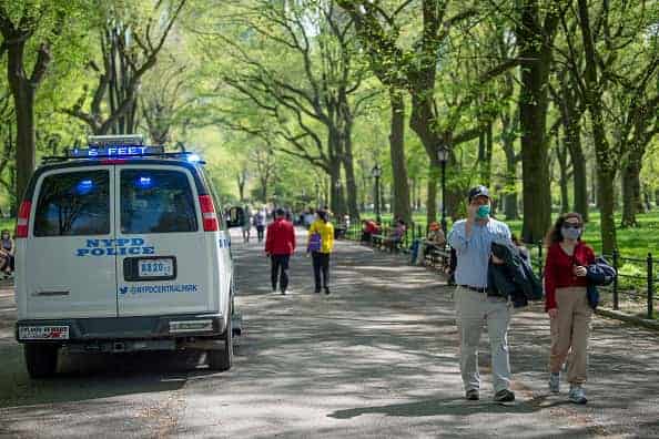 People wearing masks walk past an NYPD vehicle driving through pedestrian walkways announcing over the loud speaker to say '6 feet apart' in Central Park amid the coronavirus pandemic on May 3