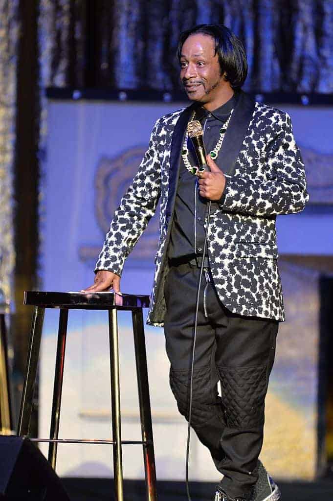 Katt Williams standing with a microphone in his hand and wearing a leopard print blazer
