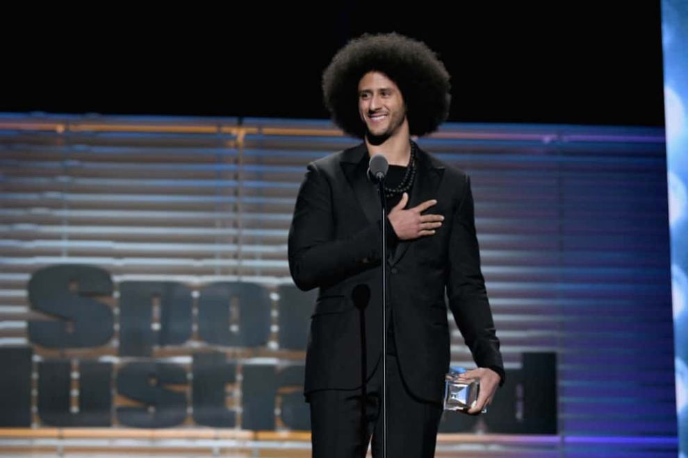 Colin Kaepernick accepting award from Sports Illustrated in all black suit with hand over heart