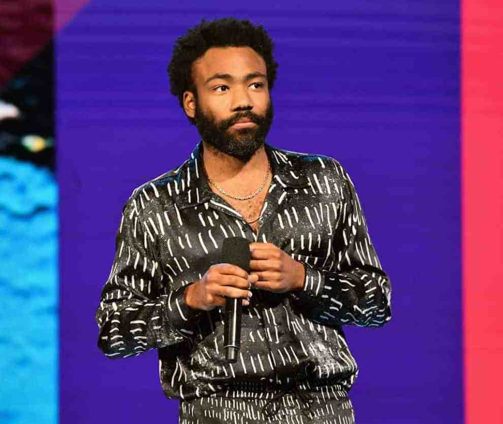 Childish Gambino wearing a black suit standing in front in a purple wall