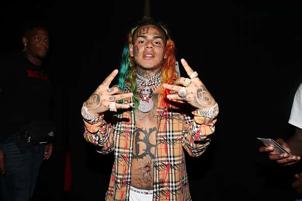 Tekashi 69 with both hands up wearing unbuttoned flannel shirt